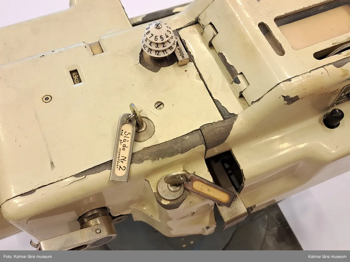 KLM 46808:12. Biljettmaskin, med tillbehör. Vändbar och vridbar maskin av metall på gråmålad kvadratisk platta, med vitmålat hölje, knappar, spakar, vridbart datumhjul och två stycken nyckellås med tillhörande nycklar. På den ena nyckelbrickan texten:  Släde nr 2 end. för reperatörer. På den andra texten: MASKIN 2. Tillverkarskylt: AEG....
Tillbehör i form av delar, biljettmagasin, biljettämnen, provbiljetter, biljettstansar.
