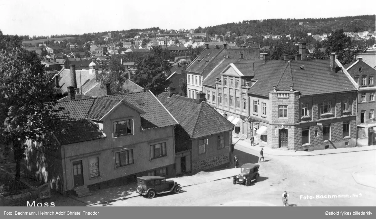 Storgata i Moss 1937-38. Hjørnet Fleischers gate og Storgata. Brehmer-gården til høyre, Storgata 16 med apotek på hjørnet. På motsatt hjørne reklame på veggen: "Carlsen &  Co A/S, Kul, koks, cinders, ved. Sundbryggene. "