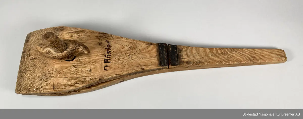 Skomakerklemme antatt laget av gran. Spadeformet redskap med klype i ene enden. Skaftet er smalt med bred kjeft. Den ene av kjeftene er fast, den andre er hengslet med en lærbit (naglet fast med trenagler), denne har gått av. Kjeftene har en svak buing på både utsiden og innsiden. For å stramme kjeftene rundt arbeidsstykket, skrues det til med en (teljet/spikket) "tremutter" med vinger. Denne mutteren har gjenger som passer til en gjenget trestang som går tvers gjennom begge kjeftene. Et stort trestykke er borte fra ene kjeften (den som sitter fast i skaftet), rett ovenfor gjennomgående hull. Består av tre deler. Svimerket med initialene O. Röstad