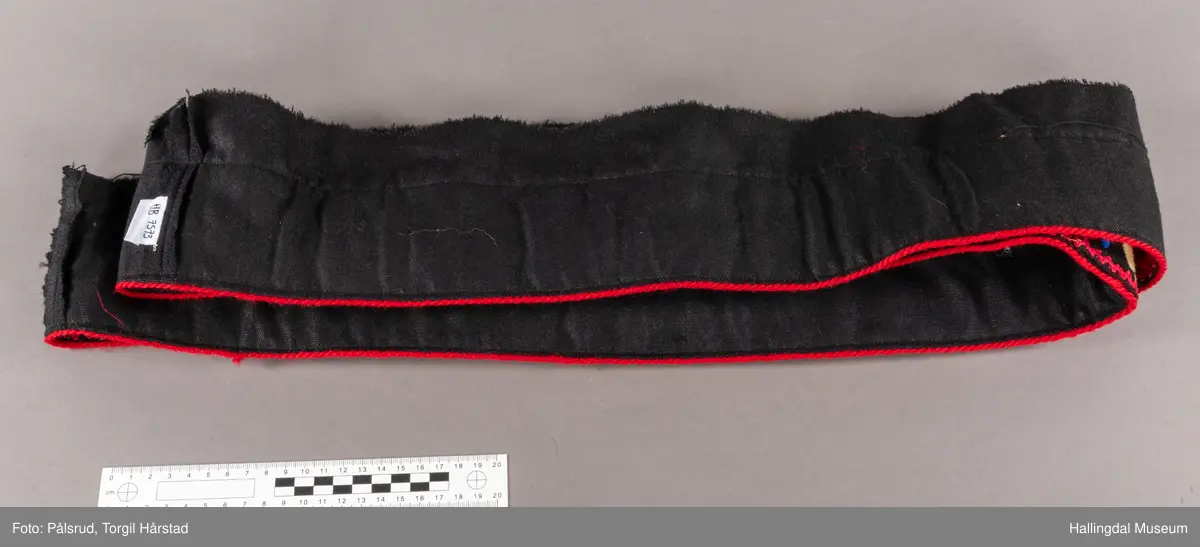 Sauma på svart klede, "gådnsauma", kanta med raude kroklisser og tvinna raud slitesnor i nedrekanten. Klypt av ein svart stakk, noko av stakken følgjer med.
