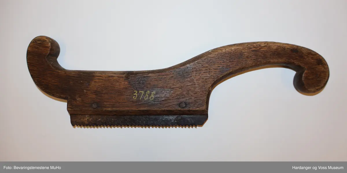 Lita handsag av tre med rektangulært, taggete sagblad av metall. Trestykket har ei avrunda form med handtak i den eine enden.