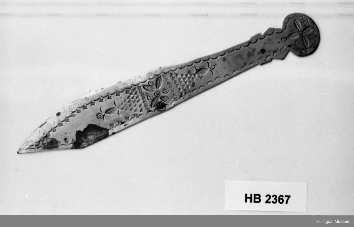 Messing, gravert dekor, blomster og ranker. Årstallet 1914 bak. L: 16 cm, bredde: 2,5 cm.