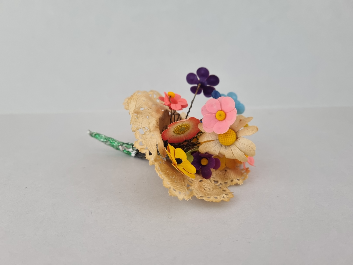 Majblommebukett i spetspapper/tårtpapper. Bunden av Beda Hallberg. Består av olika majblommor från tidigt 1907-1936. Text: Till Sylvia från Beda H.