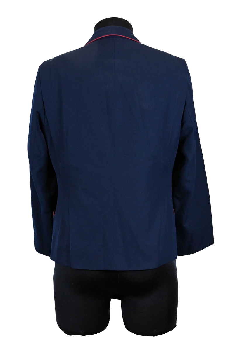 Kavaj, uniform, dammodell. Mörkblå tyg av 80% ull och 20 % polyester. Enkelknäppning med två guldfärgade metallknappar. Två midjefickor och en bröstficka på vänster sida, samt en innerficka. Kavajslaget och fickornas kanter har vinröda passpoaler. Design av Getti S.P.A.