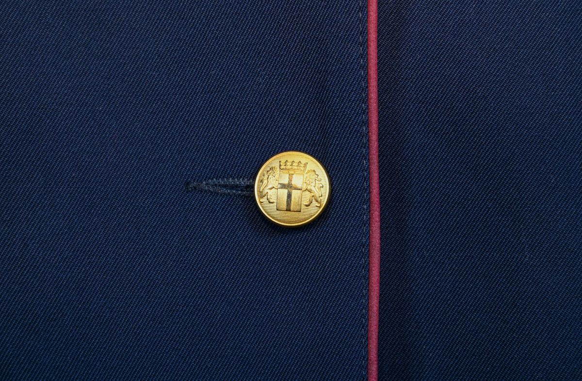 Kavaj, uniform, dammodell. Mörkblå tyg av 80% ull och 20 % polyester. Enkelknäppning med två guldfärgade metallknappar. Två midjefickor och en bröstficka på vänster sida, samt en innerficka. Kavajslaget och fickornas kanter har vinröda passpoaler. Design av Getti S.P.A.