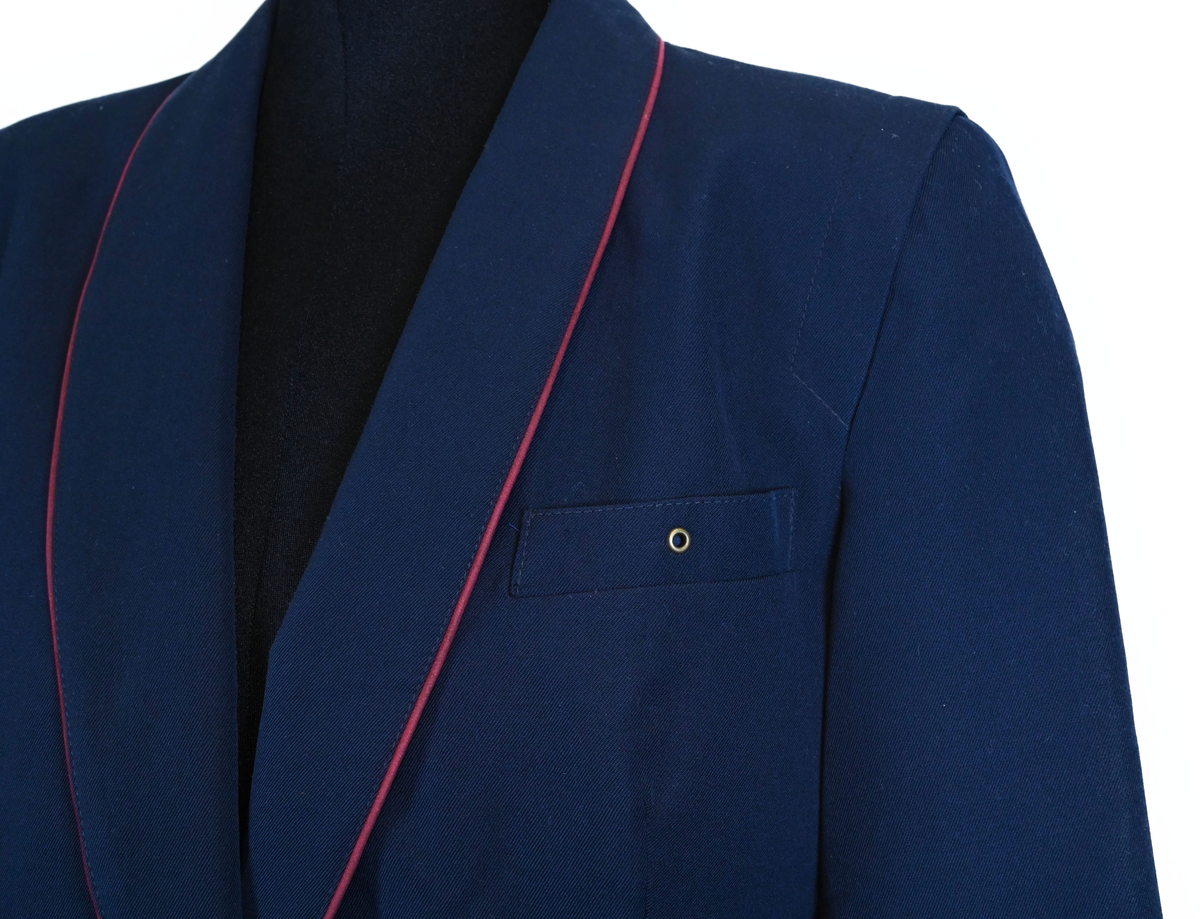 Kavaj, uniform, dammodell. Mörkblå tyg av 80% ull och 20 % polyester. Enkelknäppning med två guldfärgade metallknappar. Två midjefickor och en bröstficka på vänster sida, samt en innerficka. Kavajslaget och fickornas kanter har vinröda passpoaler. Design av Getti S.P.A.