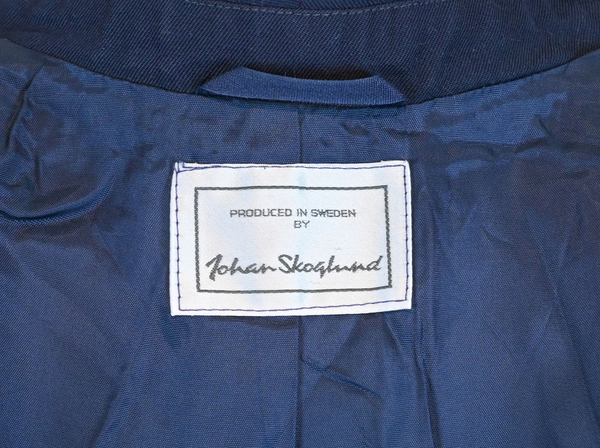 Kavaj, uniform, dammodell. Mörkblå tyg av 80% ull och 20 % polyester. Enkelknäppning med två guldfärgade metallknappar. Två midjefickor och en bröstficka på vänster sida, samt en innerficka. Kavajslaget och fickornas kanter har vinröda passpoaler. Design av Getti S.P.A.