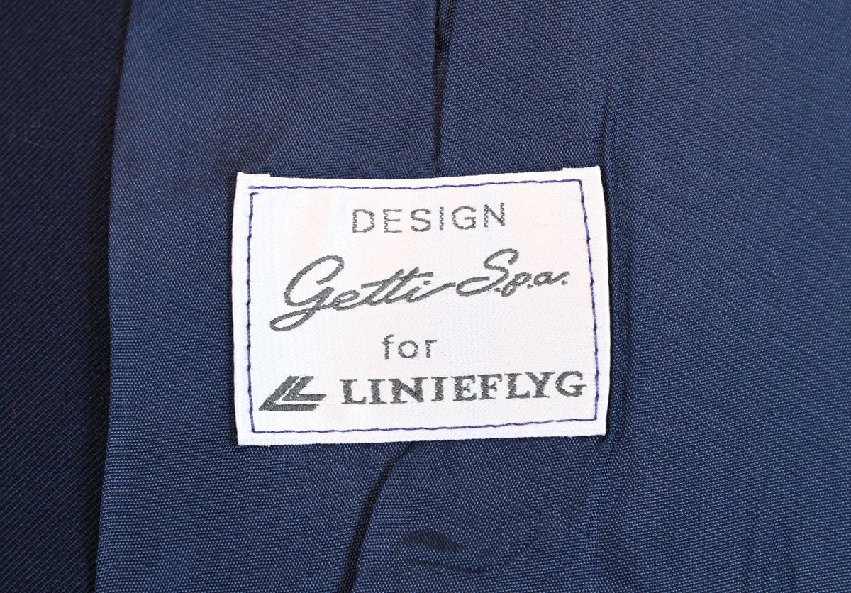 Kavaj, uniform, dammodell. Mörkblå tyg av 80% ull och 20 % polyester. Enkelknäppning med två guldfärgade metallknappar. Två midjefickor och en bröstficka på vänster sida, samt en innerficka. Kavajslaget och fickornas kanter har vinröda passpoaler. Design av Getti S.P.A.