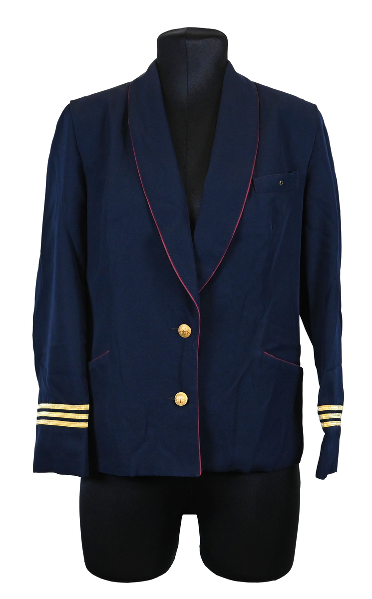 Kavaj, uniform, dammodell. Mörkblå tyg av 80% ull och 20 % polyester. Enkelkläppning med två guldfärgade knappar och tre rader av guldgaloner vid vardera ärmslut. Två midjefickor och en bröstficka på vänster sida. Kavajslaget och fickornas kanter har vinröda passpoaler. Design av Getti S.P.A.