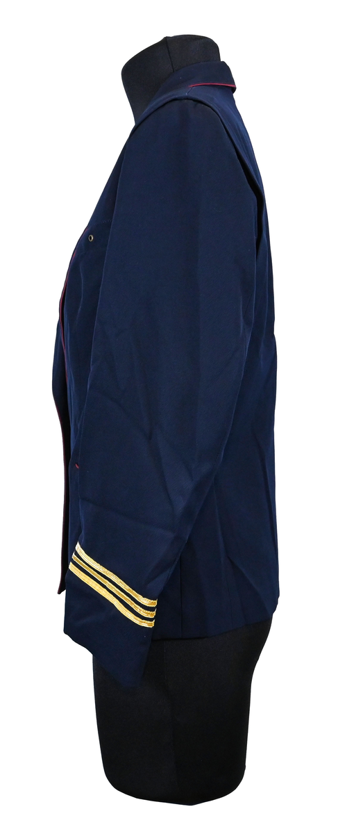 Kavaj, uniform, dammodell. Mörkblå tyg av 80% ull och 20 % polyester. Enkelkläppning med två guldfärgade knappar och tre rader av guldgaloner vid vardera ärmslut. Två midjefickor och en bröstficka på vänster sida. Kavajslaget och fickornas kanter har vinröda passpoaler. Design av Getti S.P.A.