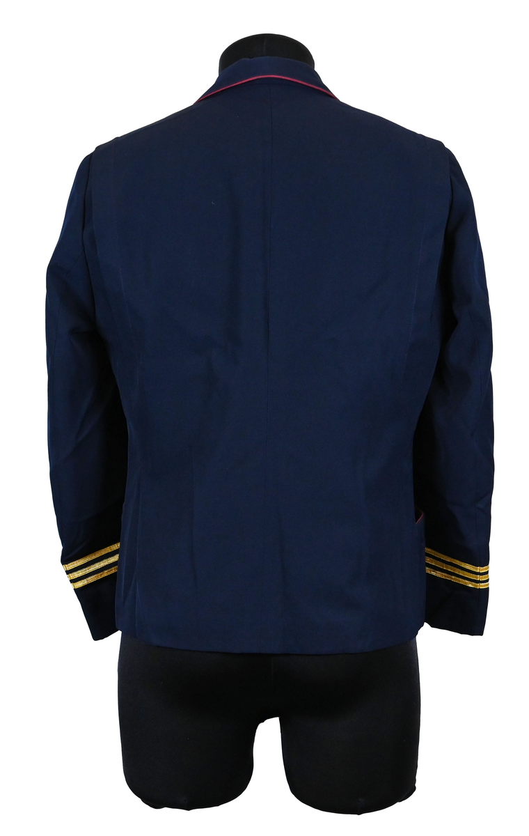 Kavaj, uniform, dammodell. Mörkblå tyg av 80% ull och 20 % polyester. Enkelkläppning med två guldfärgade knappar och tre rader av guldgaloner vid vardera ärmslut. Två midjefickor och en bröstficka på vänster sida. Kavajslaget och fickornas kanter har vinröda passpoaler. Design av Getti S.P.A.