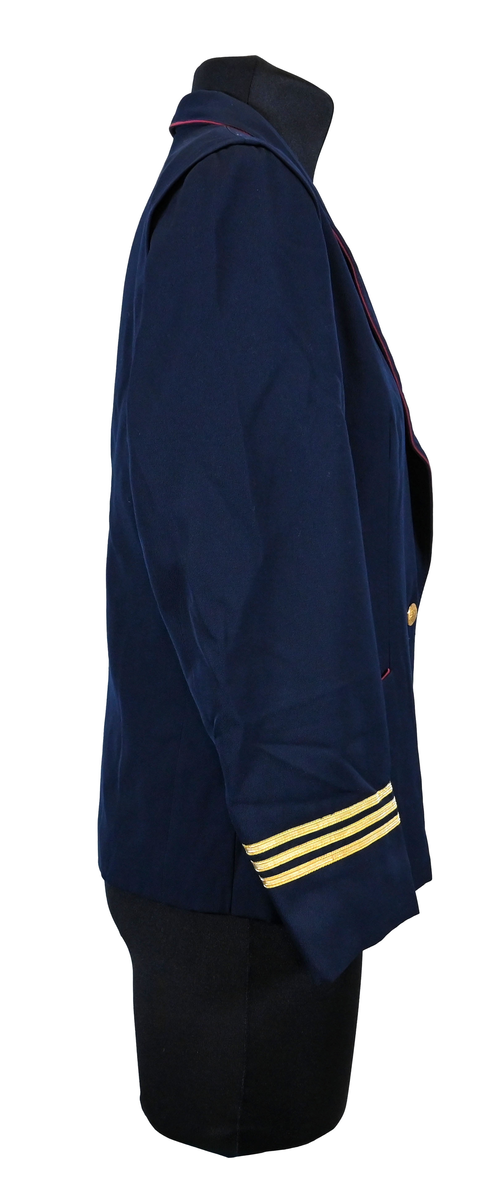 Kavaj, uniform, dammodell. Mörkblå tyg av 80% ull och 20 % polyester. Enkelkläppning med två guldfärgade knappar och tre rader av guldgaloner vid vardera ärmslut. Två midjefickor och en bröstficka på vänster sida. Kavajslaget och fickornas kanter har vinröda passpoaler. Design av Getti S.P.A.