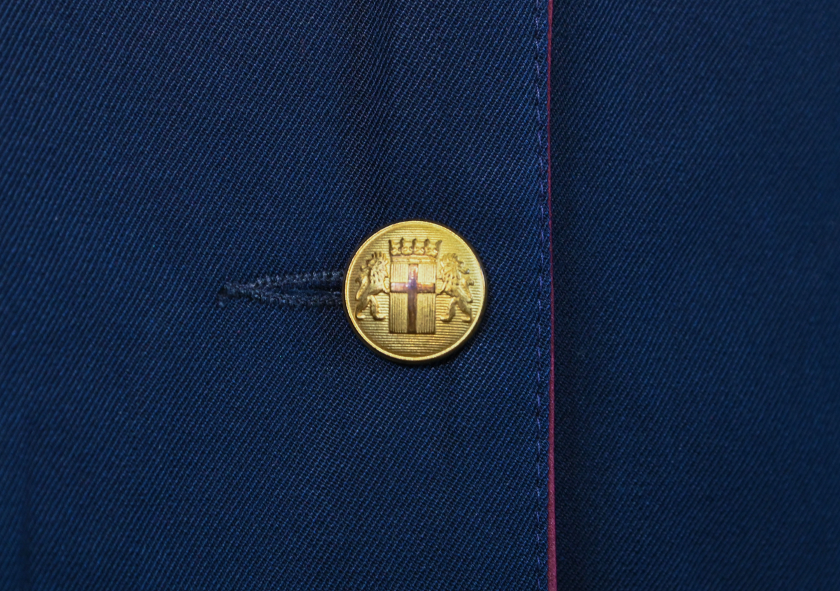 Kavaj, uniform, dammodell. Mörkblå tyg av 80% ull och 20 % polyester. Enkelkläppning med två guldfärgade knappar och tre rader av guldgaloner vid vardera ärmslut. Två midjefickor och en bröstficka på vänster sida. Kavajslaget och fickornas kanter har vinröda passpoaler. Design av Getti S.P.A.