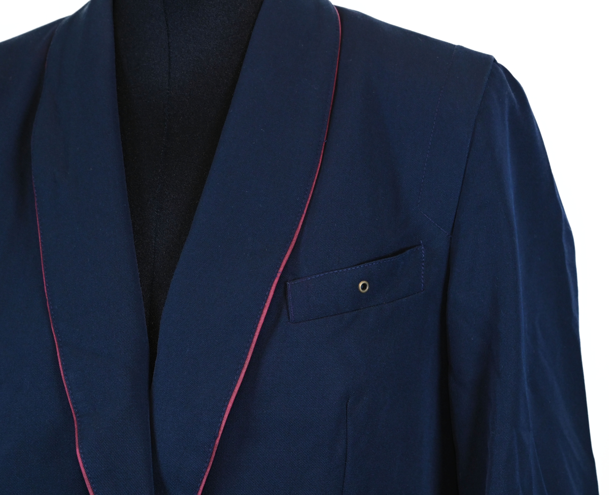 Kavaj, uniform, dammodell. Mörkblå tyg av 80% ull och 20 % polyester. Enkelkläppning med två guldfärgade knappar och tre rader av guldgaloner vid vardera ärmslut. Två midjefickor och en bröstficka på vänster sida. Kavajslaget och fickornas kanter har vinröda passpoaler. Design av Getti S.P.A.