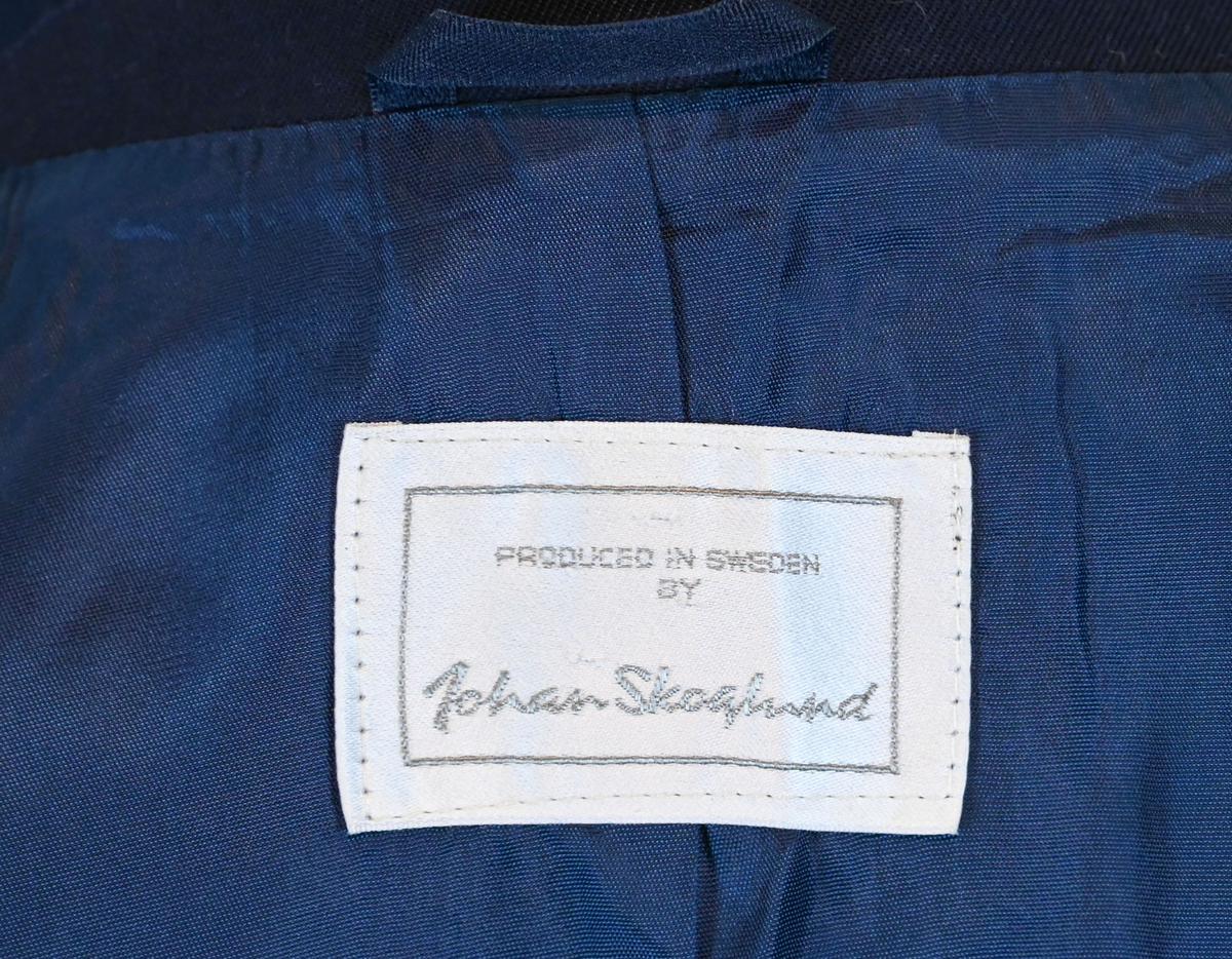 Kavaj, uniform, dammodell. Mörkblå tyg av 80% ull och 20 % polyester. Enkelkläppning med två guldfärgade knappar och tre rader av guldgaloner vid vardera ärmslut. Två midjefickor och en bröstficka på vänster sida. Kavajslaget och fickornas kanter har vinröda passpoaler. Design av Getti S.P.A.