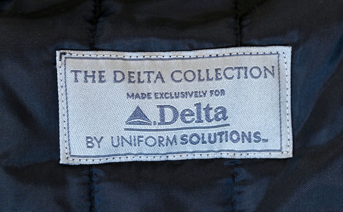 Fodrad överrock i trenchcoat-modell för flygbolagspersonal hos Delta Air Lines. Rocken har två ytterfickor samt en innerficka. Rocken består av skalrock samt löstagbart foder genom dragkedja. Både skalrock och foder är svarta och skalrocken är sydd med raglanärm. Enkel knäppning med fem svarta plastknappar samt två svarta plastknappar vid vardera ärmslut.

Storlek 42 R