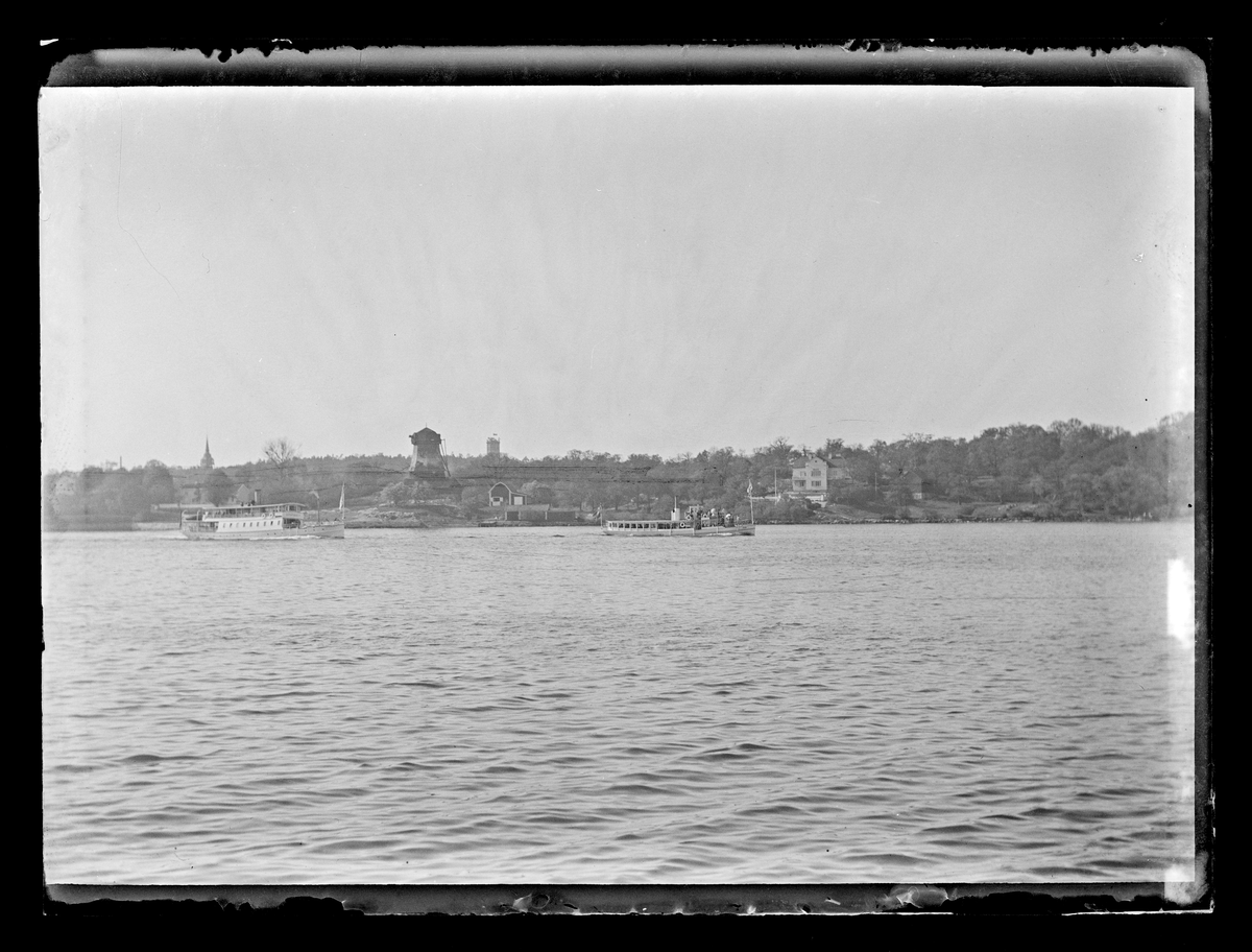 Bilden visar kusten av Waldemarsudde i Stockholm med ett kyrktorn, väderkvarn och flera villor längs strandkanten.