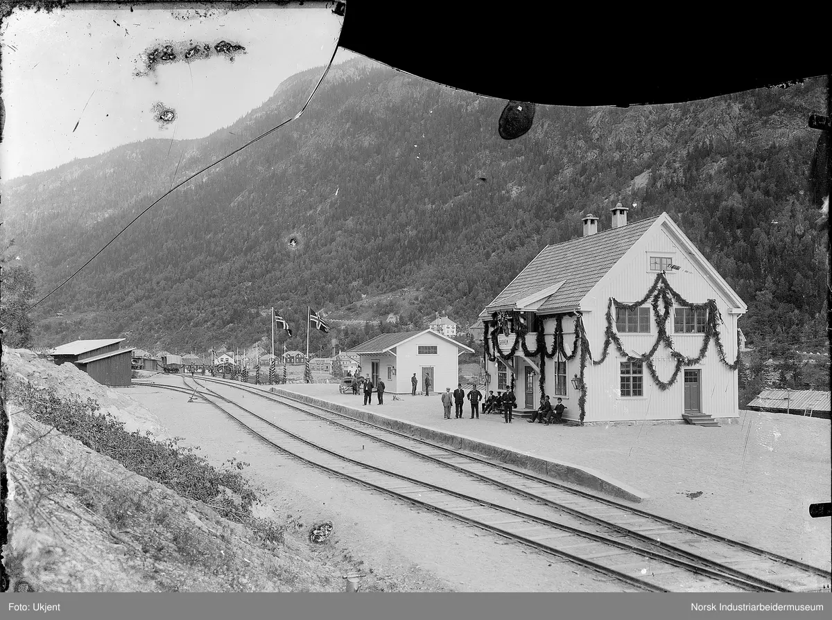 Åpningen av Rjukan Jernbanestasjon. Bygningen er pyntet med girlander.  Mennesker er samlet på perrongen, hest og kjerre i bakgrunnen. To flaggstenger med det norske flagg heist. Boliger i Villaveien og Flekkebyen i bakgrunnen.