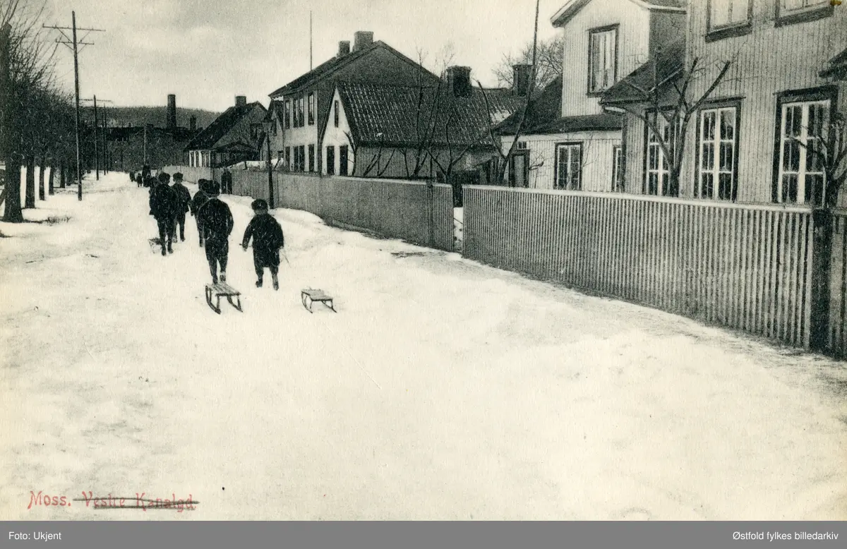 Gateparti fra Strandgata i Moss vinterstid. Tittelen på postkortet er "Moss, Vestre Kanalgate", gatenavn er overstrøket. Barn med kjelker går i gata.