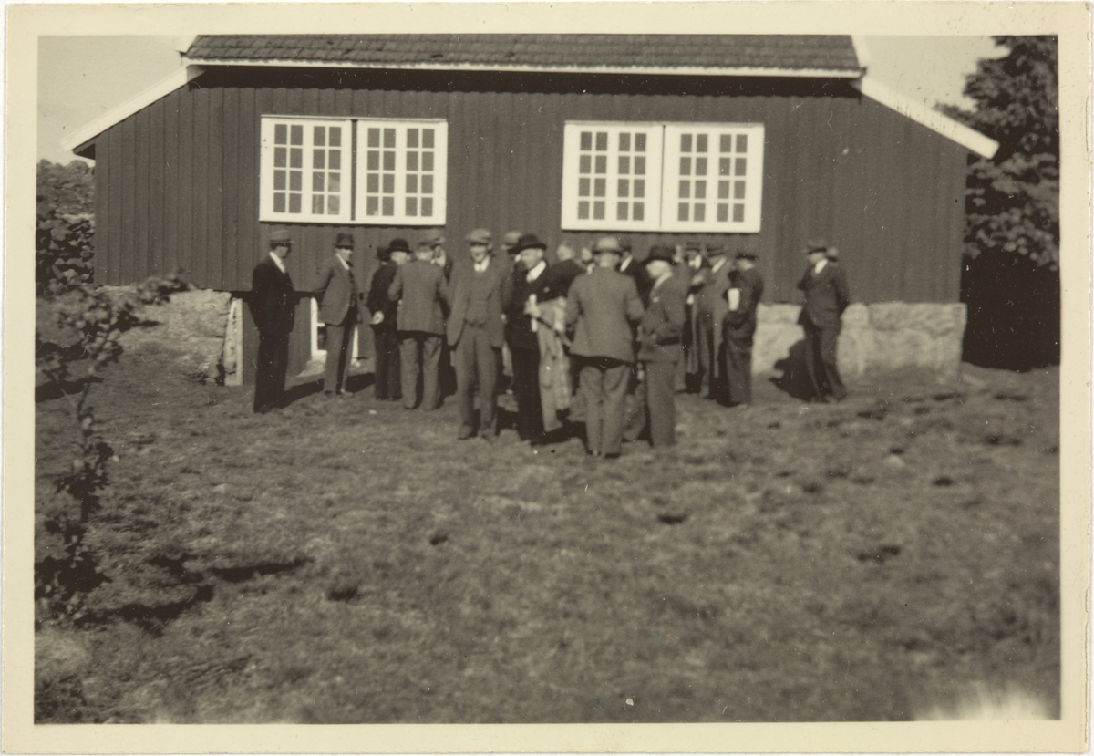 Ei gruppe menn står framføre eit hus: Knudaheio. Samling med folk ute i hagen.