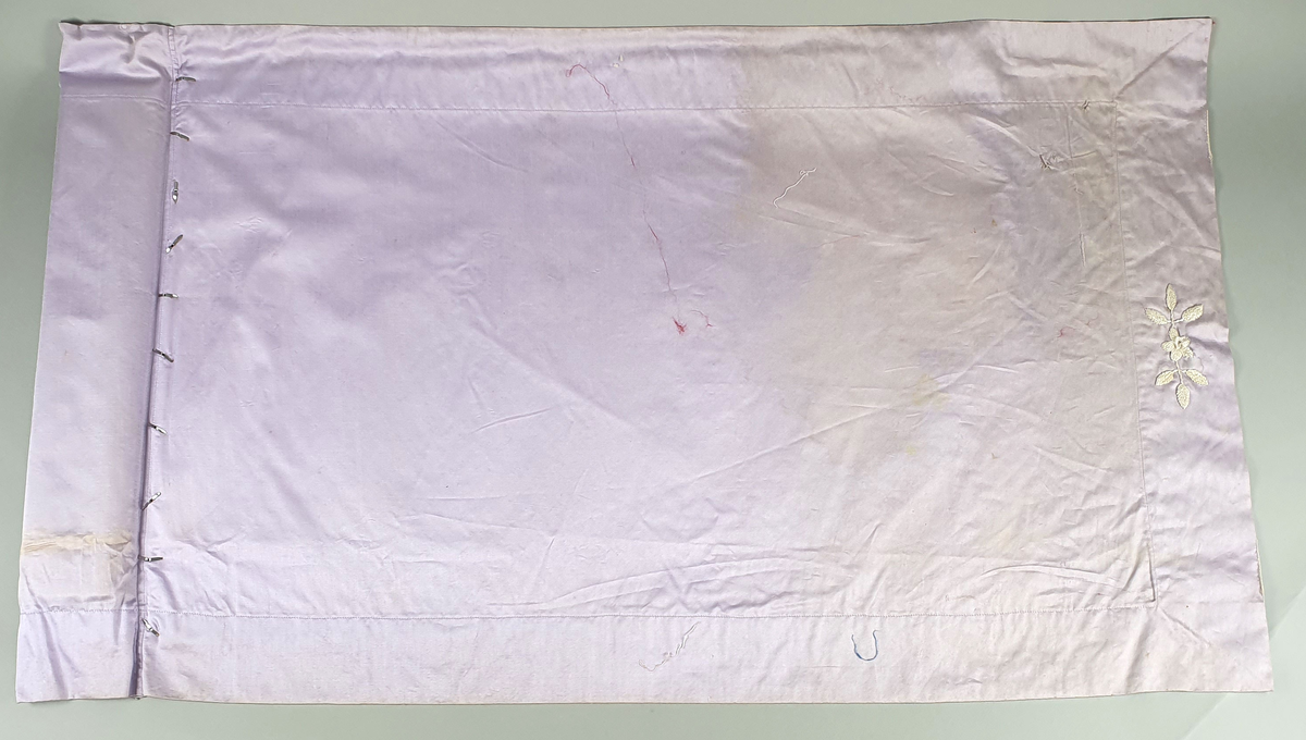Mappe av lys lilla bomull med en brodert hvit blomst på den ene kortenden. På den ene siden er det festet 7 tynne silkepapir. I mappen føger 6 håndbroderte duker i ulike fasonger og mønstre. 
A: Rund duk med heklet bord, av bomull
B: Oval duk med røde broderte liljer, av bomull
C: Rund duk med røde broderte blomster og blad, av lin
D: Rund duk med heklet blondekant, av lin
E: Brikke med røde, broderte bokstaver og tall, av strie
F: Rund duk med brodert bladkrans i hvitsøm, av bomull