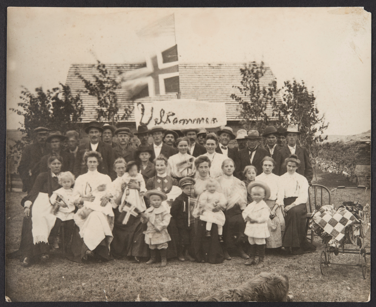 Stort  gruppebilde av mennesker med det norske og det amerikanske flagg og en plakat som sier "Velkommen" i bakgrunnen. Skrevet på baksiden av bildet: J. J. Gilseth, Mohall, N. Dak.