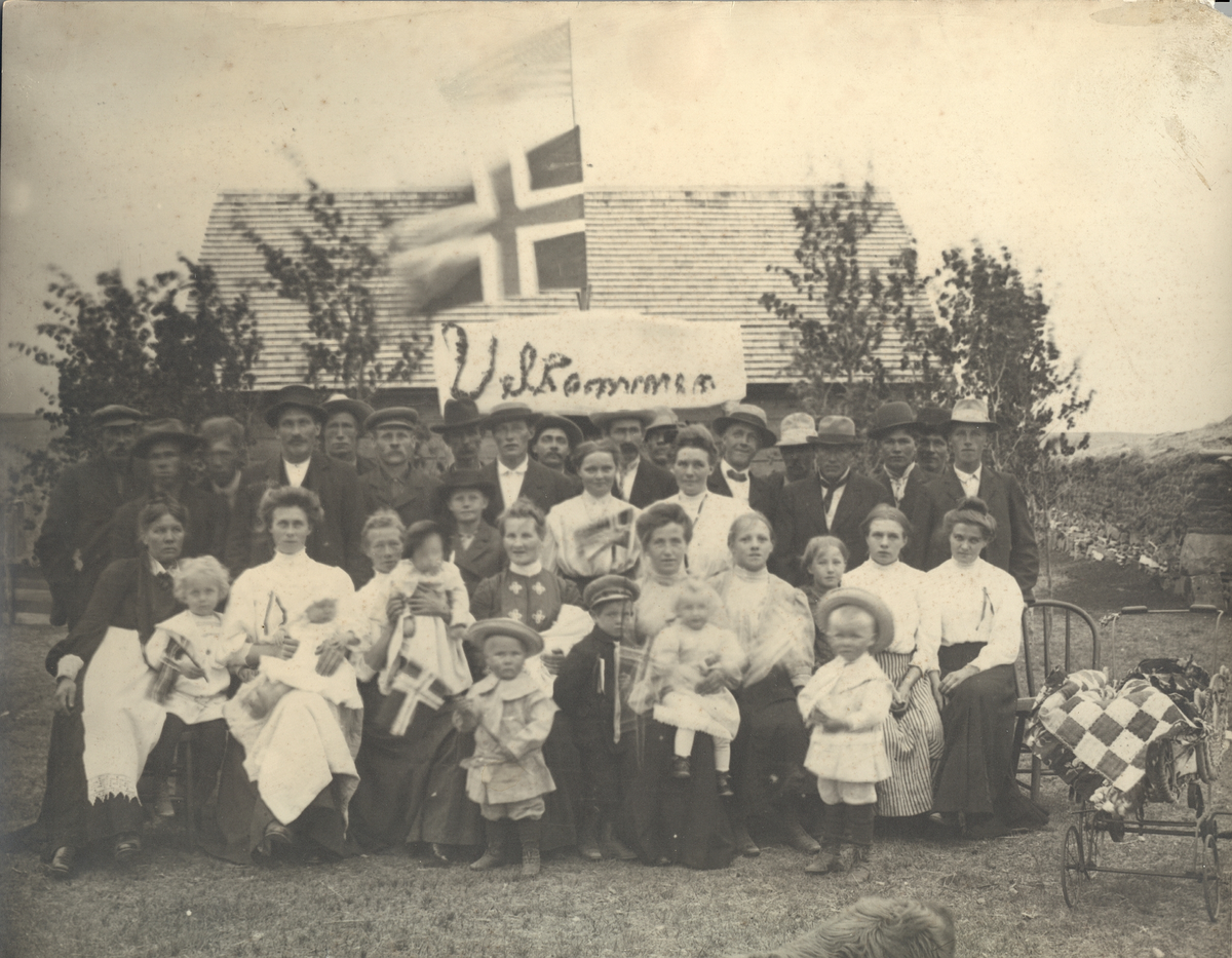 Stort  gruppebilde av mennesker med det norske og det amerikanske flagg og en plakat som sier "Velkommen" i bakgrunnen. Skrevet på baksiden av bildet: J. J. Gilseth, Mohall, N. Dak.