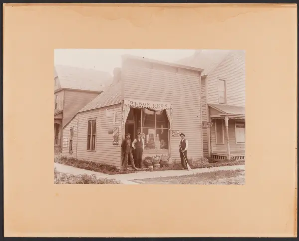 Butikk "Olson Bros", tre menn utenfor, - den ene med feiekos