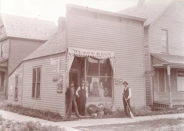 Butikk "Olson Bros", tre menn utenfor, - den ene med feiekos