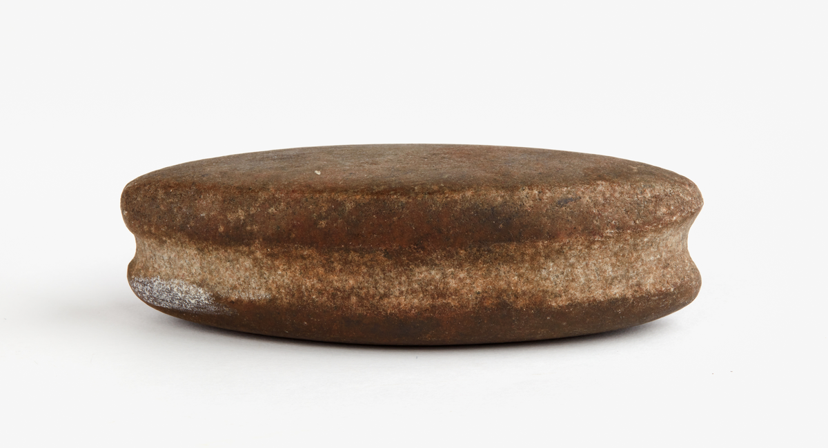 Eldslagningssten av mörkbrun kvartsit med spetsoval form. En 12 mm bred och grund ränna runt om. Ett smalt slagspår på ena sidan. Slipad, utom rännan längst smalsidorna.