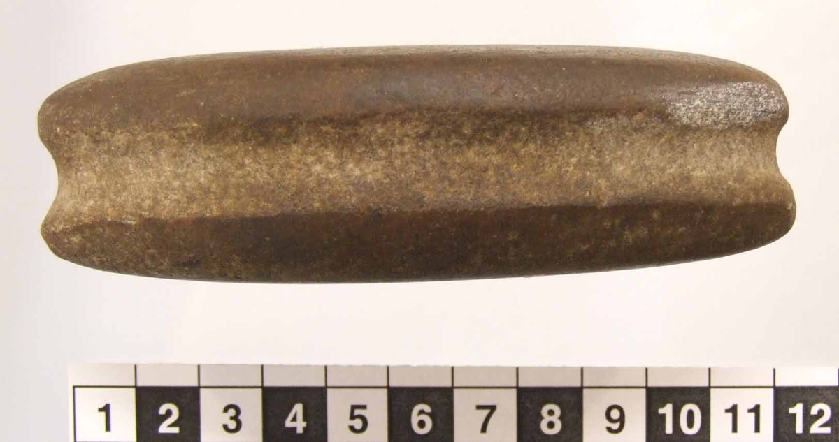 Eldslagningssten av mörkbrun kvartsit med spetsoval form. En 12 mm bred och grund ränna runt om. Ett smalt slagspår på ena sidan. Slipad, utom rännan längst smalsidorna.