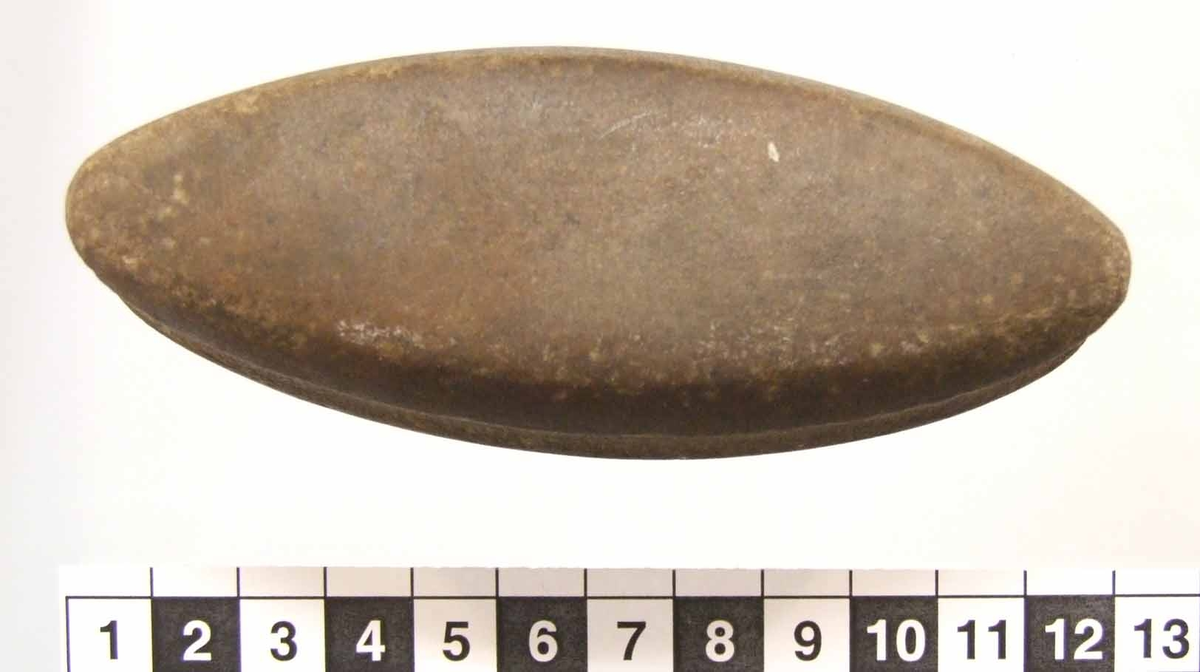 Eldslagningssten av mörkbrun kvartsit med spetsoval form. En 12 mm bred och grund ränna runt om. Ett smalt slagspår på ena sidan. Slipad, utom rännan längst smalsidorna.