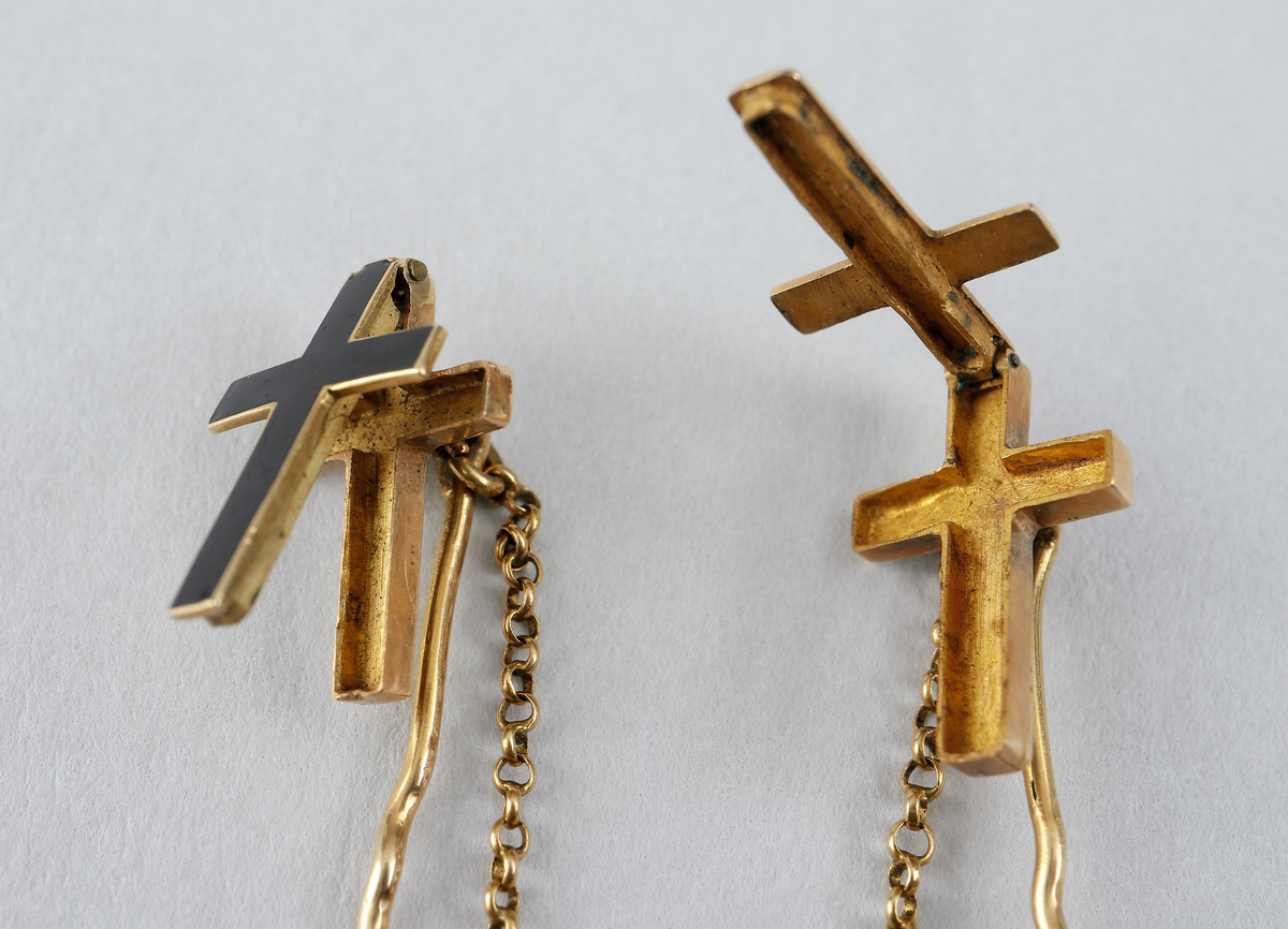 Två kors, nålförsedda med en kedja emellan. Bägge korsens framsidor går att fälla ut, och innanför finns en liten gömma. Smycket är tillverkat i 18 karat guld med svartfärgade framsidor. Smycket förvaras i en röd pappask med oläslig text, dansk, och ej tillhörande. Stämplat: 18K tre kronor (kattfot) IES G4.