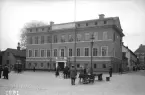 Linköping 1939 och några av tidens människor har gjort sig ärende till Stora torgets södra delar. Det gamla rådhuset i Linköping var som sådant en kompromiss som samlat flera parallella funktioner. Domprostboställe, polisstation och bank var några verksamheter som rådmännen hade att dela huset med. Fotoåret hade magistratens förutsättning i huset dock förbättrats.