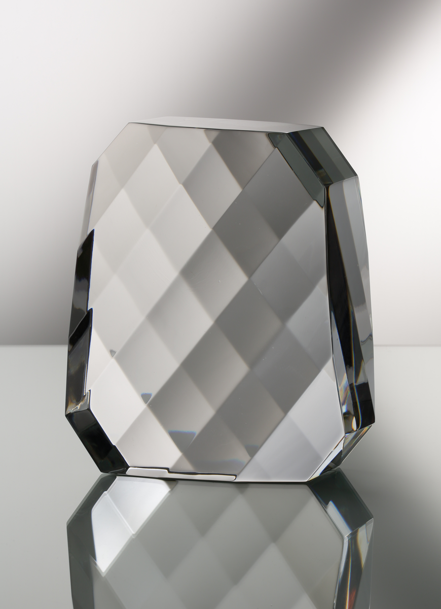 Formgivare: Jan Johansson. Skulptur i massivt klarglas. Rektangulär form med brutna hörn. Sju slipade diagonala fasetter på främre och bakre vägg som bildar ett "visuellt rutmönster".