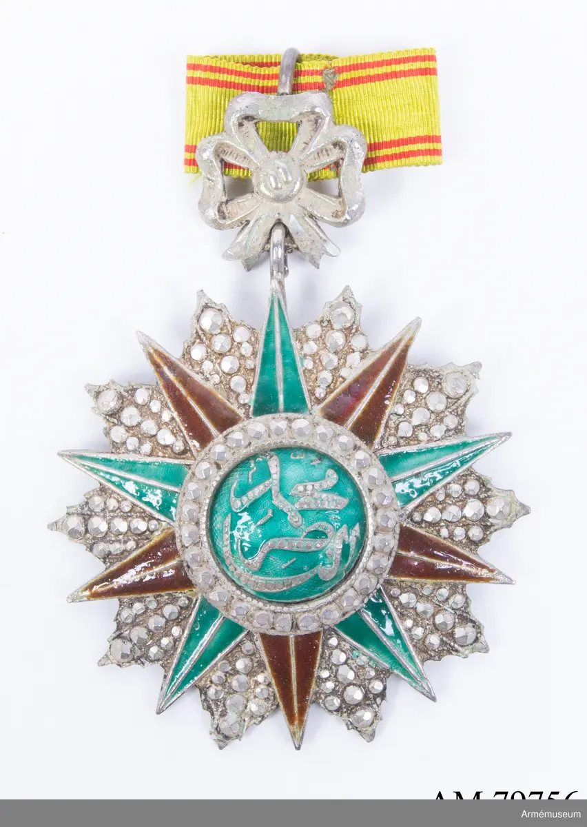 Ordenstecken för kommendör.

En tiouddig stjärna (varannan spets av röd, varannan av grön emalj, varje spets längs mitten avdelad av en silverrand); stjärnan är lagd på en tiouddig facetterad silverkraschan.

Ovanpå stjärnan en rund sköld, omgiven av en ring med briljantslipade silverfacetter. Skölden är av grön emalj, och å densamma läses i silverslingor den regerande beyens  namnchiffer.

Ordenstecknet är fäst vid en silverrosett.

Bandet, ljust mossgrönt, har två smala, röda ränder på vardera sidan. Flera lösa samhörande band.
//Kompl. beskr. PRN