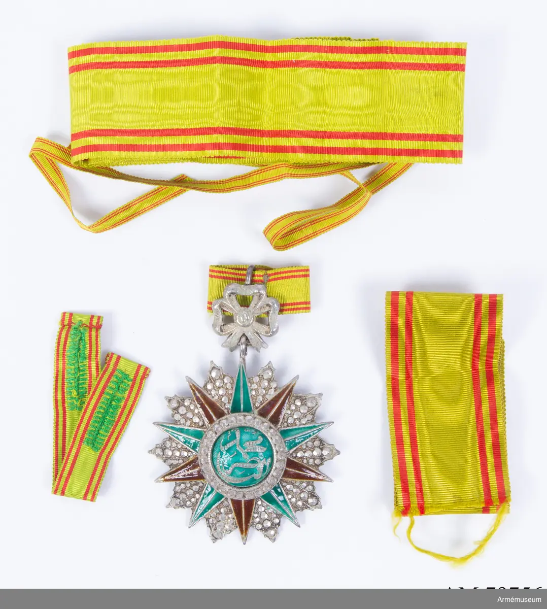 Ordenstecken för kommendör.

En tiouddig stjärna (varannan spets av röd, varannan av grön emalj, varje spets längs mitten avdelad av en silverrand); stjärnan är lagd på en tiouddig facetterad silverkraschan.

Ovanpå stjärnan en rund sköld, omgiven av en ring med briljantslipade silverfacetter. Skölden är av grön emalj, och å densamma läses i silverslingor den regerande beyens  namnchiffer.

Ordenstecknet är fäst vid en silverrosett.

Bandet, ljust mossgrönt, har två smala, röda ränder på vardera sidan. Flera lösa samhörande band.
//Kompl. beskr. PRN