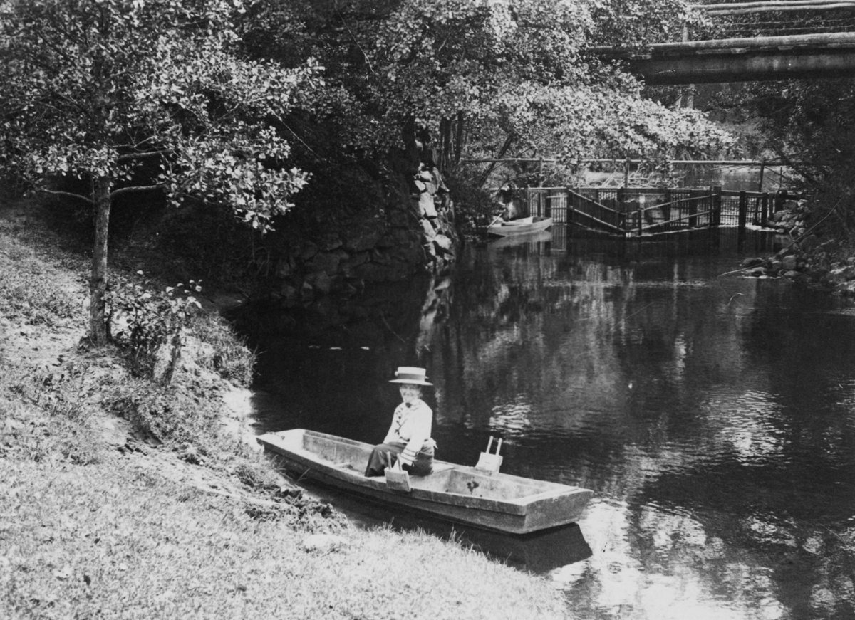 "Laxgård och hängbro i Uddaveka på 1920-talet." I förgrunden sitter en kvinna i stor hatt i en eka vid åstranden.