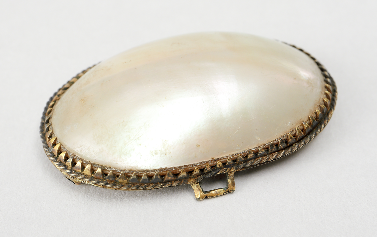 Ovalt spänne (för dam) av pärlemo, infattat i förgylld metall. Infattningen är gjord i uddmönster. Undersidans ram har två längsgående rör, samt en hake fastlödda.

Inskrivet i huvudbok 1867.