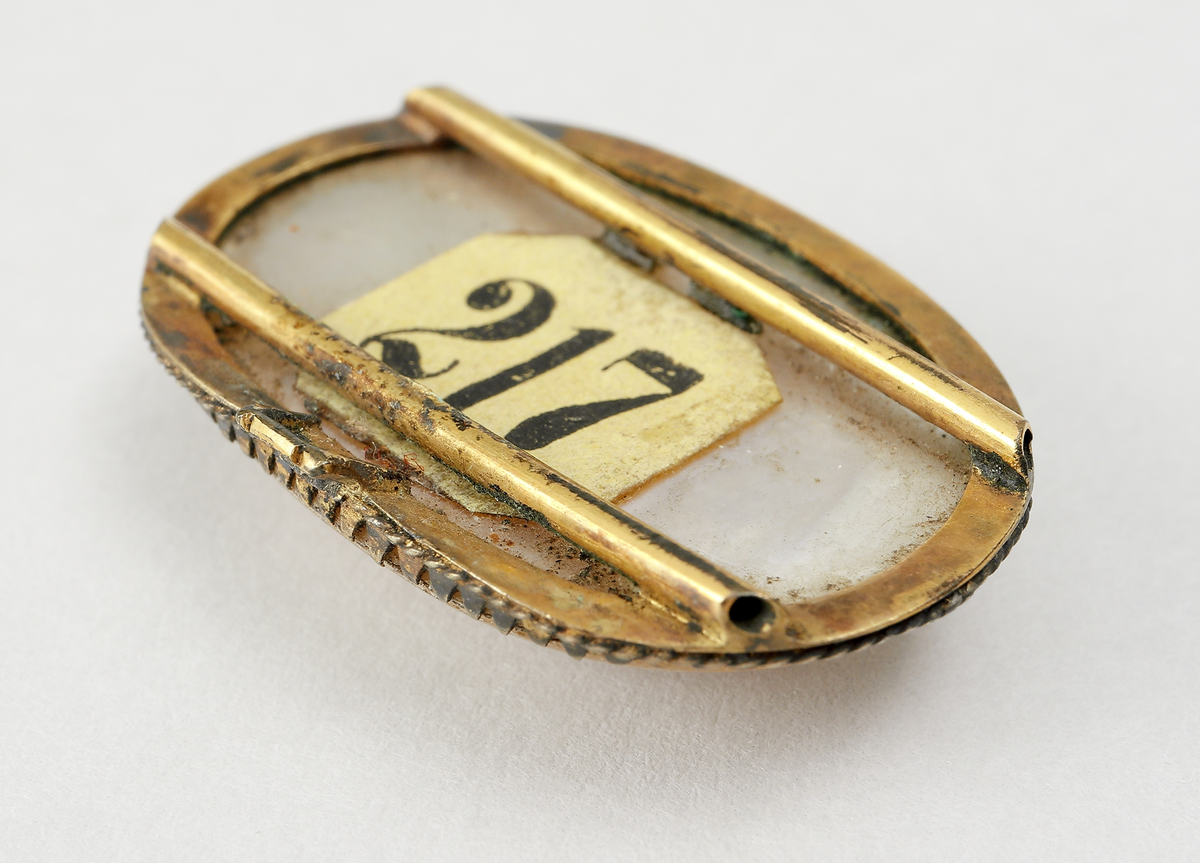 Ovalt spänne (för dam) av pärlemo, infattat i förgylld metall. Infattningen är gjord i uddmönster. Undersidans ram har två längsgående rör, samt en hake fastlödda.

Inskrivet i huvudbok 1867.