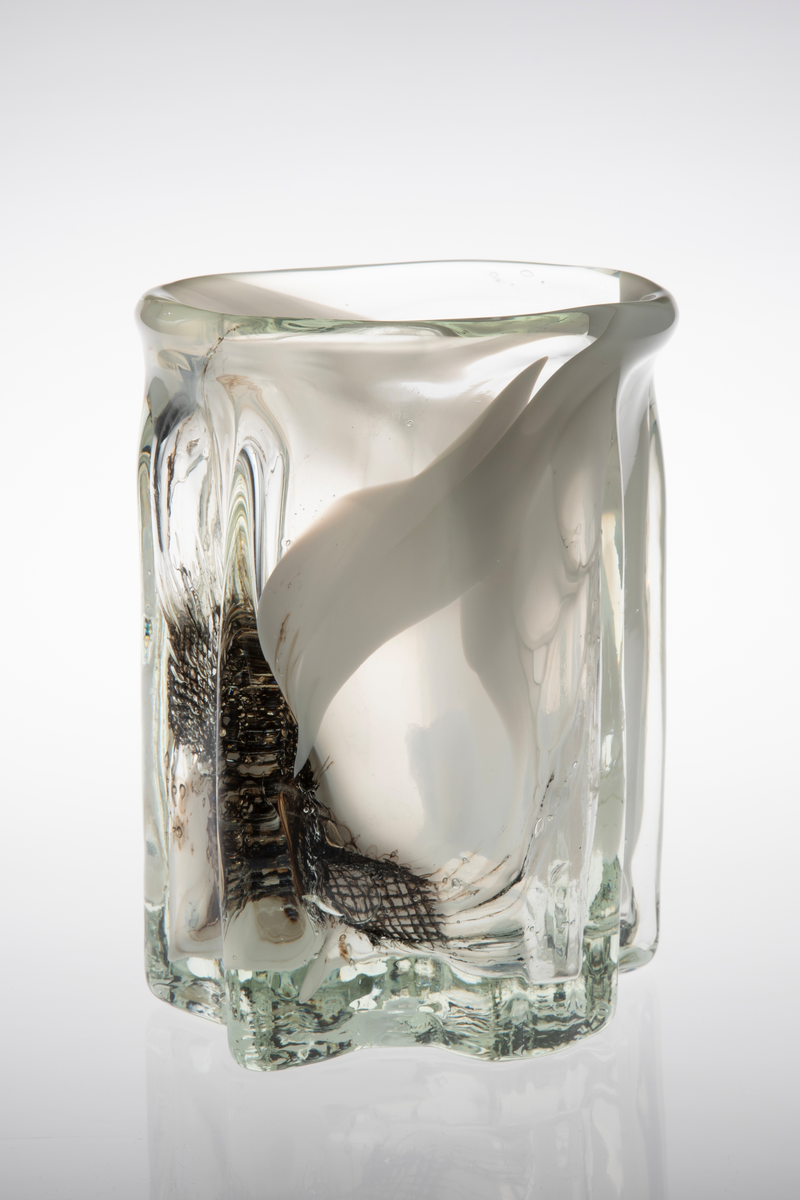 Tilnærmet sylindrisk vase i klart glass med innlagt dekor. Vasens tykke vegger har bølgende former og avsluttes i en sirkulær irregulær munningsrand. Innslag av melkehvite partier i opalinglass, samt fiberduk og luftblærer.