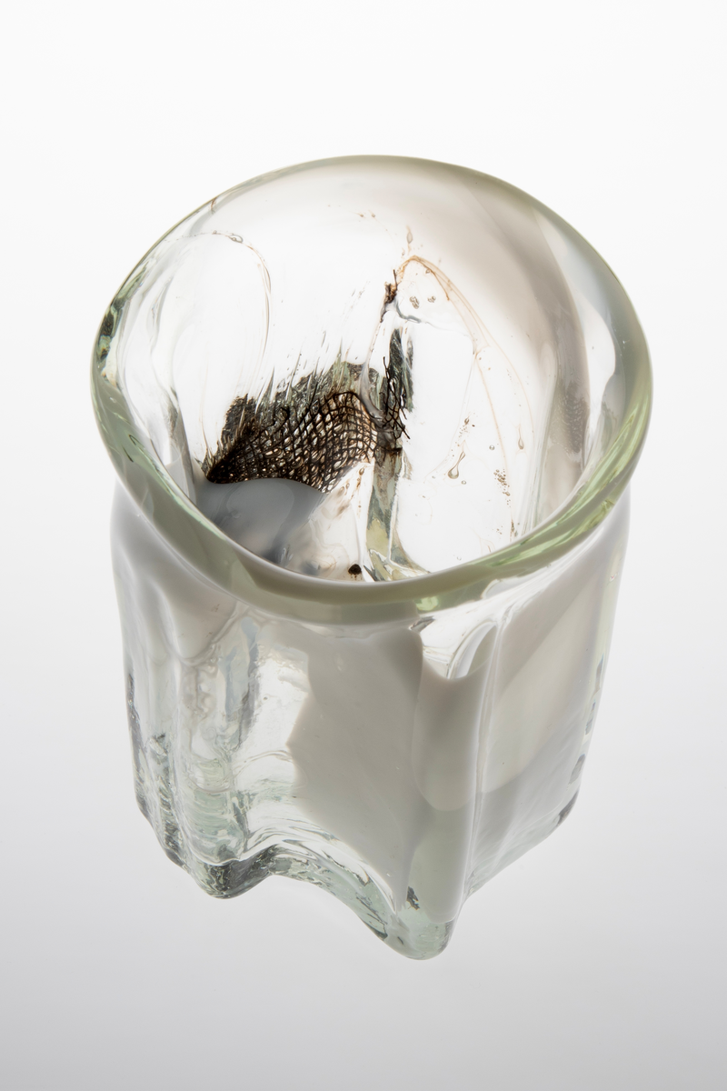 Tilnærmet sylindrisk vase i klart glass med innlagt dekor. Vasens tykke vegger har bølgende former og avsluttes i en sirkulær irregulær munningsrand. Innslag av melkehvite partier i opalinglass, samt fiberduk og luftblærer.