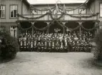 Gruppfoto av en stor samling officerare och civila herrar i frack och andra ämbetsuniformer, avfotograferade framför den festsmyckade officersmässen på Kronobergs hed. 
Källa: Svensk Rikskalender 1908,s. 173.