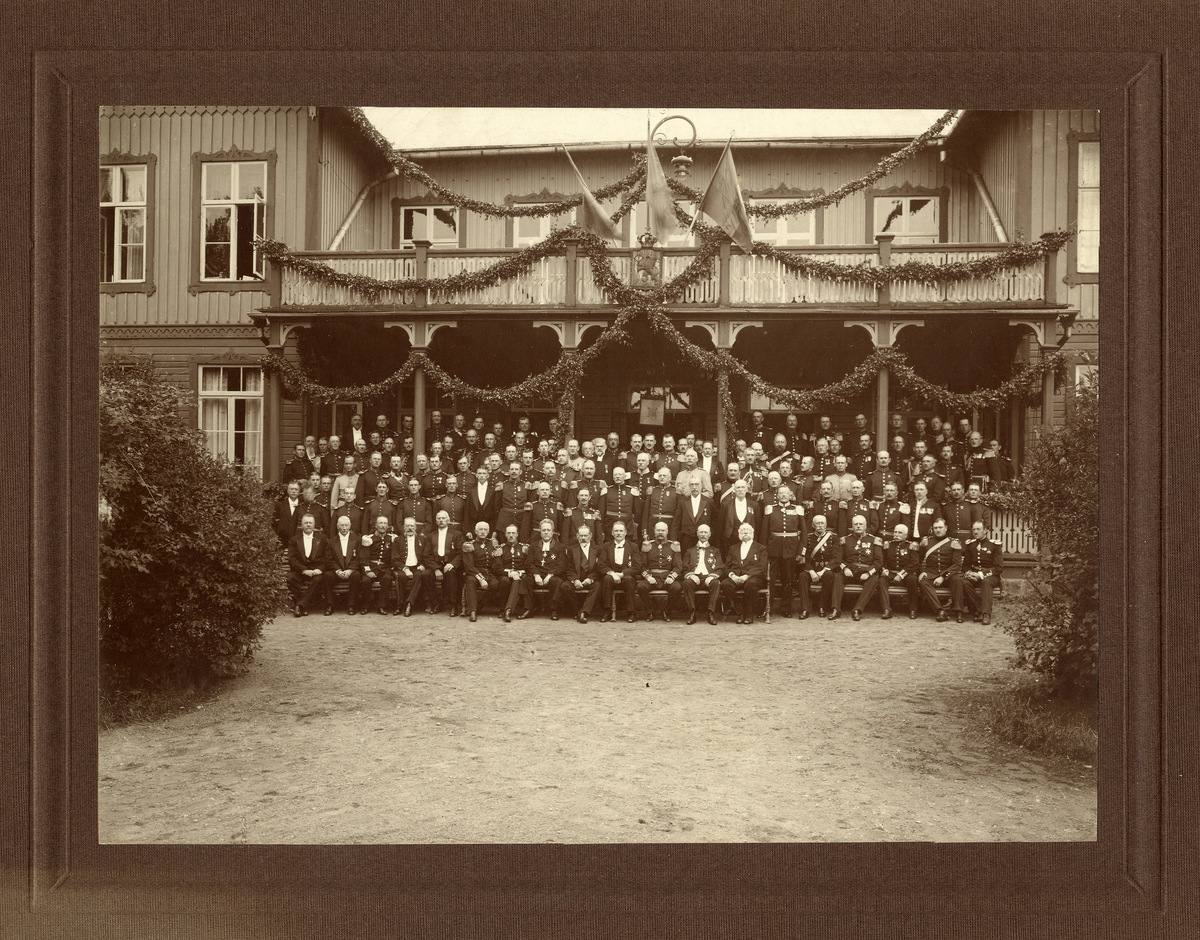 Gruppfoto av en stor samling officerare och civila herrar i frack och andra ämbetsuniformer, avfotograferade framför den festsmyckade officersmässen på Kronobergs hed. 
Källa: Svensk Rikskalender 1908,s. 173.