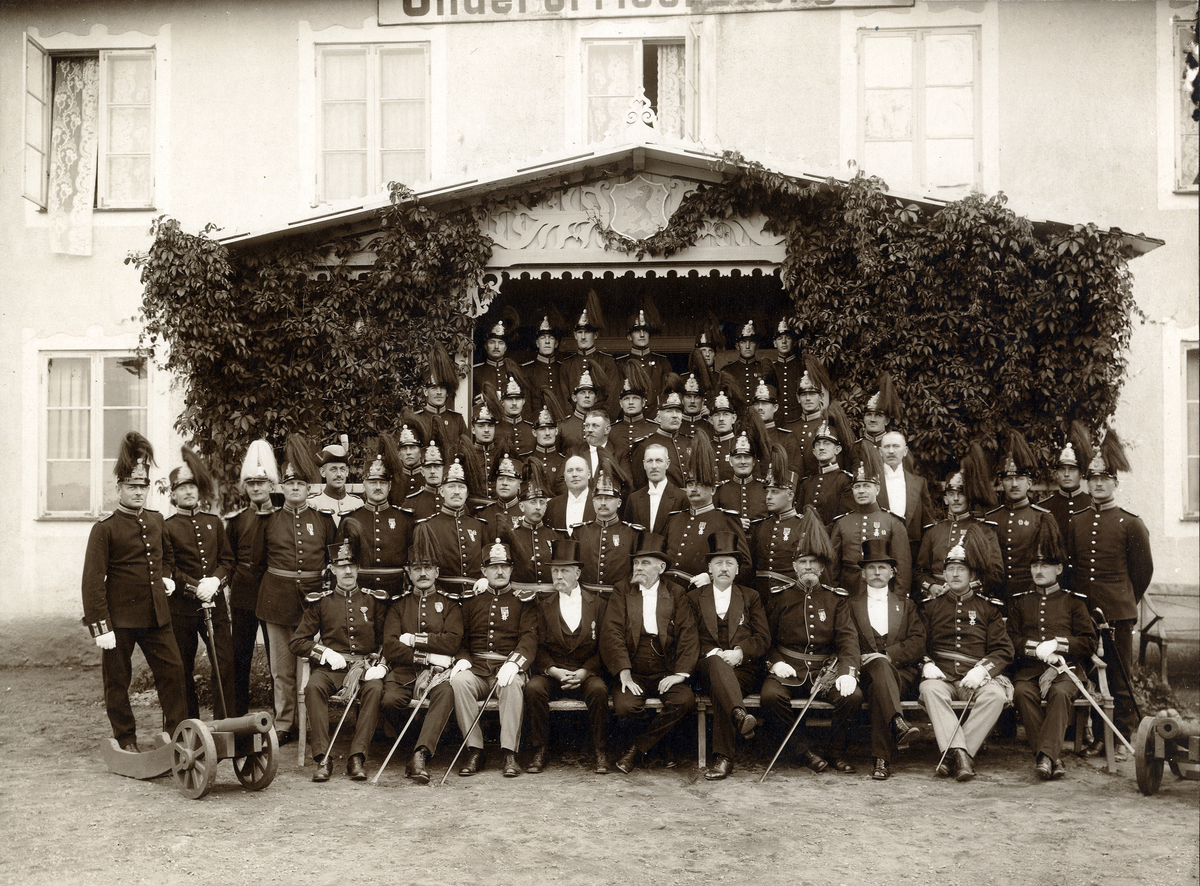 Gruppfoto av ett antal underofficerare och andra, flertalet i paraduniform med kask, men också en del i frack med hög hatt.
De är fotograferade framför verandan till ett större, reveterat hus. 
Källa: Svensk Rikskalender 1908,s. 173.
