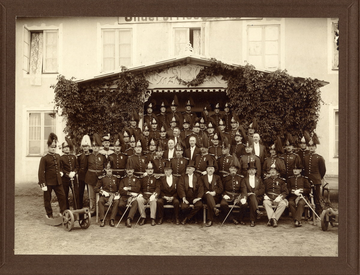 Gruppfoto av ett antal underofficerare och andra, flertalet i paraduniform med kask, men också en del i frack med hög hatt.
De är fotograferade framför verandan till ett större, reveterat hus. 
Källa: Svensk Rikskalender 1908,s. 173.