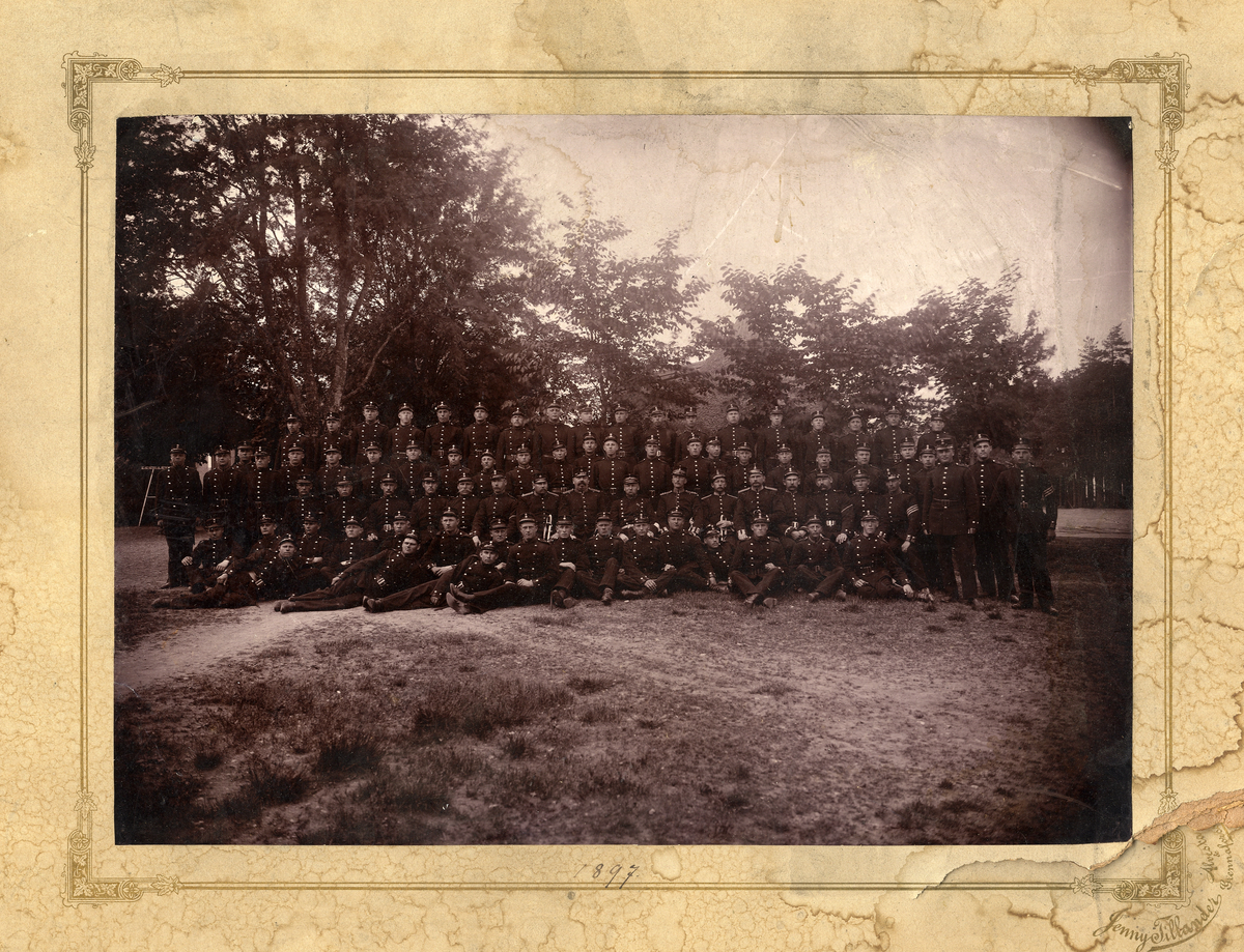 Gruppfoto av ett antal meniga/rekryter med sina officerare, uppställda på en grusplan framför en dunge.