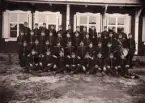 Gruppfoto av ett antal meniga/rekryter, uppställda utomhus. 
I mitten sitter några officerare. Till höger syns en regementsmusiker med diverse olika instrument.
