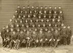 Gruppfoto av ett antal meniga/rekryter, uppställda utomhus, framför en rödmålad byggnad.
I mitten sitter några officerare.