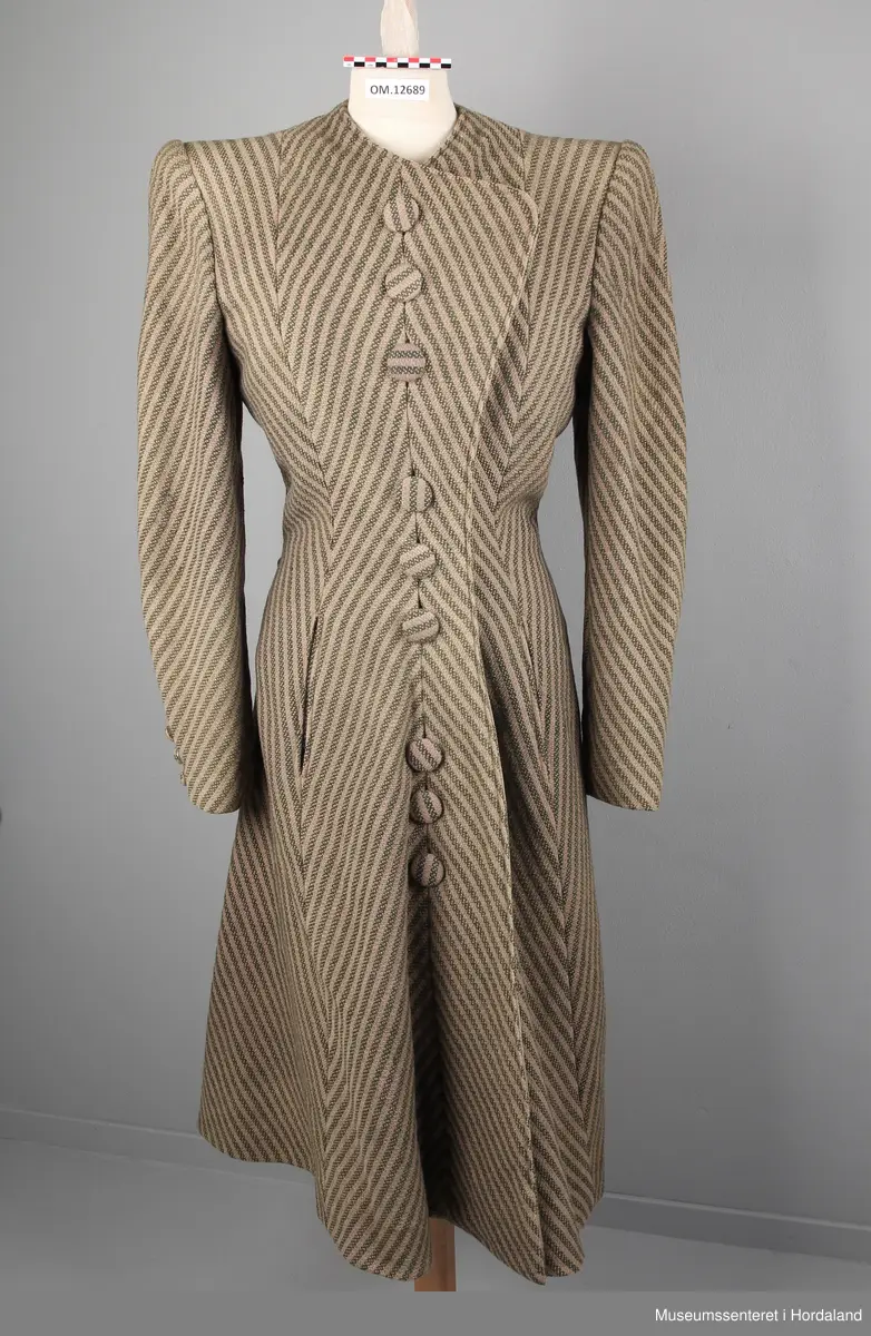 Kåpa er sydd saman av 10 bredder (skråklipte), der alle stripene møtes rett i alle saumane. Fasongsydd og sterkt innsvinga i livet. Slag, men ikkje krage. Trekte knappar - 9 stk i same stoff som kåpa. Knapphola ligg i ein saum - altså ikkje klipte opp og sydde. To stikklommer på kvar side framme i saumen. Tre trekte knappar framme på ermene. Fora med kunstsilkestoff. På innsida nede har kåpa skoning (forsterking). Store skulderputer.